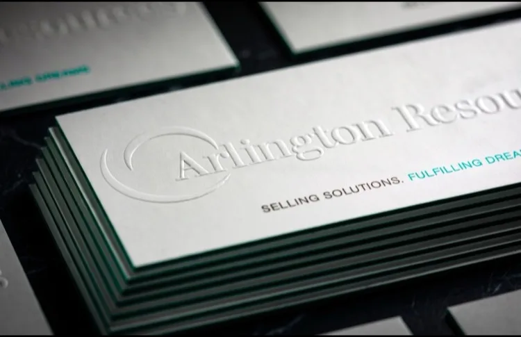 A macro close-up of a white "Arlington Resources" business card with a blind-embossed logo and a teal foiled edge, the raised relief catching the light.