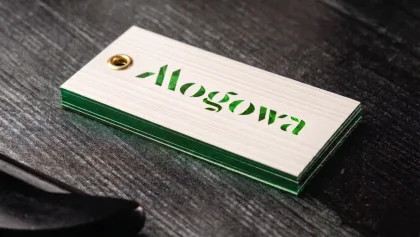 A natural cotton hang tag with a brass eyelet and a deep green embossed 'Alogowa' wordmark, showing a foiled green edge, on a dark wood surface.