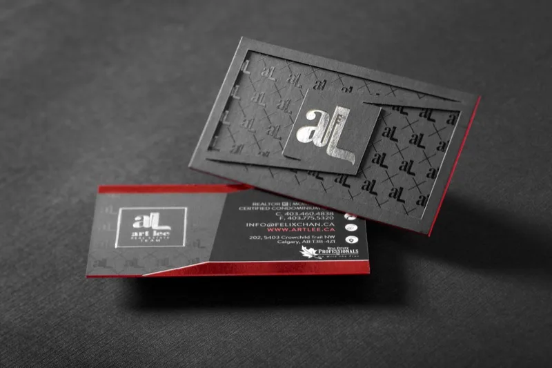 A multi-board dark gray real-estate business card with a silver foil monogram and red colored edges, standing upright beside the closed card.