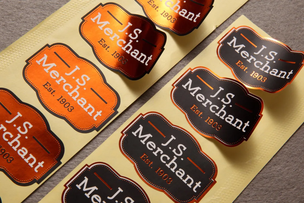 A printed roll of vinyl label stickers in copper and charcoal colorways, die-cut to a scalloped badge shape and ready to peel.